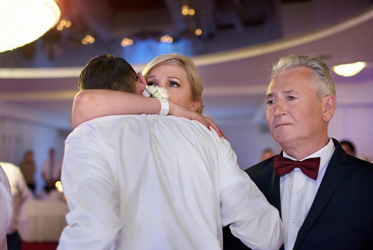 Wzruszona panna młoda w objęciach mężczyzny, z bukiecikiem na nadgarstku, obok stoi elegancki mężczyzna w muszce na tle sali weselnej.