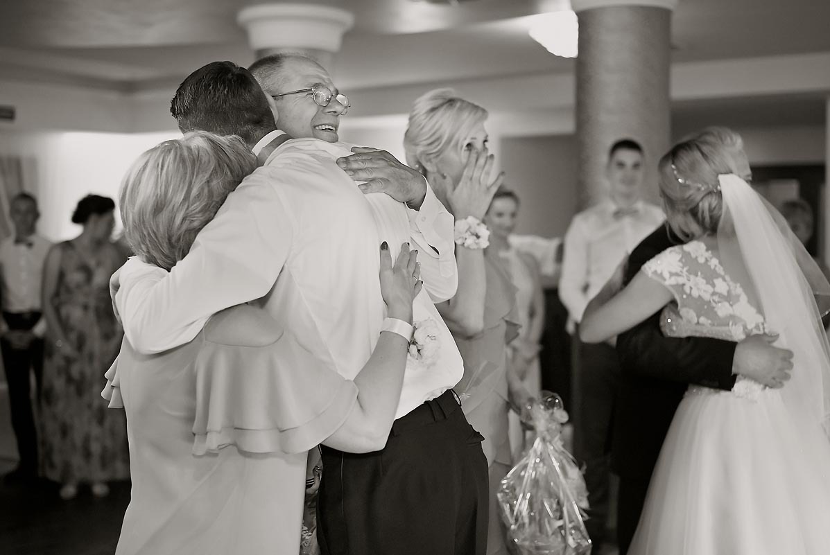 Czarno-białe zdjęcie z uroczystości ślubnej, przedstawiające wzruszonego ojca w okularach, obejmującego pannę młodą w białej sukni i welonie, w tle widoczni inni goście.