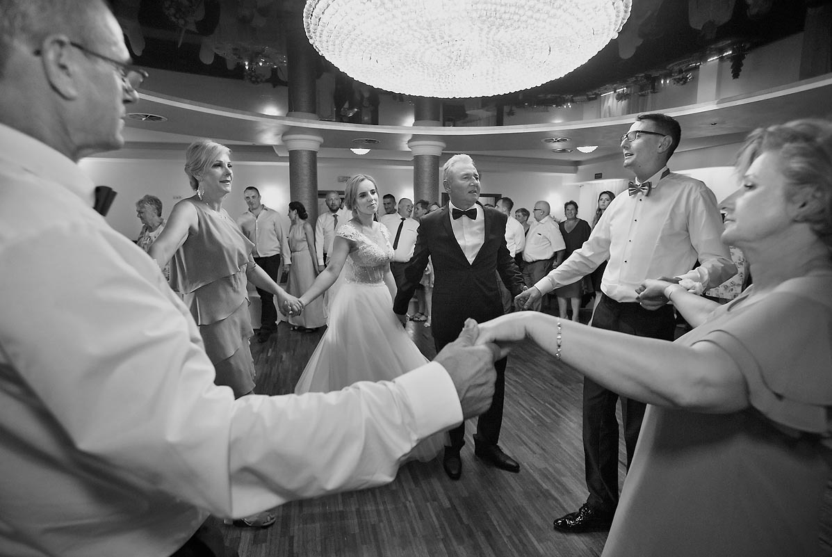 Czarno-białe zdjęcie z wesela, przedstawiające tradycyjny taniec w kręgu wokół pary młodej. Wszyscy trzymają się za ręce, a na twarzach gości widać radość. W tle widoczny żyrandol i kolumny.