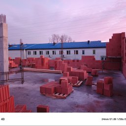 Zbigniew Jacher - Firma Remontowo-Budowlana - Wczesny etap budowy z czerwonej ceramiki poryzowanej: widoczne palety z bloczkami, fragmenty wzniesionych ścian oraz zbrojony słup na tle budynku z niebieskim dachem i różowego nieba.