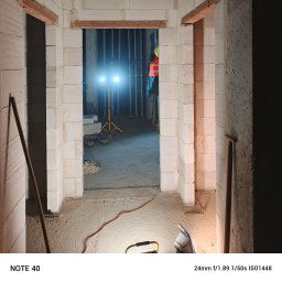 Zbigniew Jacher - Firma Remontowo-Budowlana - Wnętrze w trakcie budowy z widocznymi ścianami z białych bloczków, otwór drzwiowy z nadprożem z cegieł, oświetlony halogenem budowlanym, narzędzia budowlane oparte o ścianę.