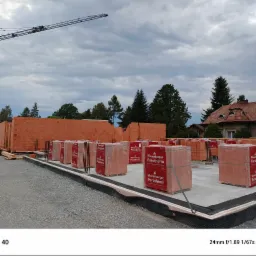 Budowa domu jednorodzinnego z czerwonej cegły Porotherm, widoczne palety z cegłami na fundamencie, w tle las i niebo z chmurami, żuraw budowlany w oddali.