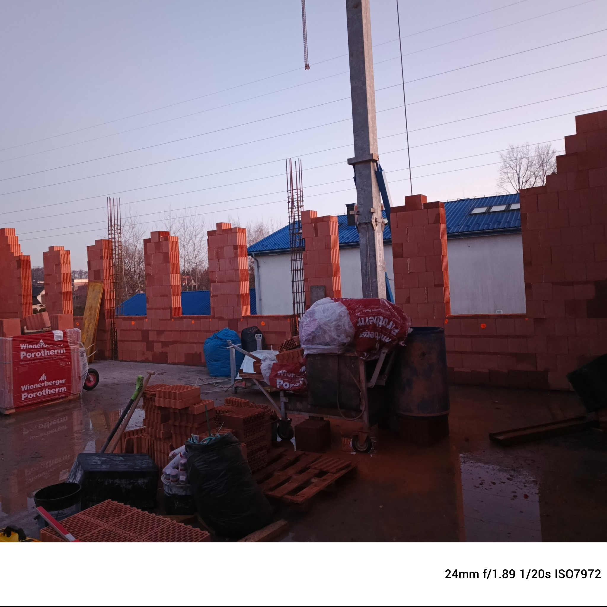 Wznoszenie murów z czerwonej cegły Porotherm w Rzezawie, widoczne palety z materiałem, rusztowanie i częściowo wymurowane ściany z otworami okiennymi na tle budynku z niebieskim dachem.