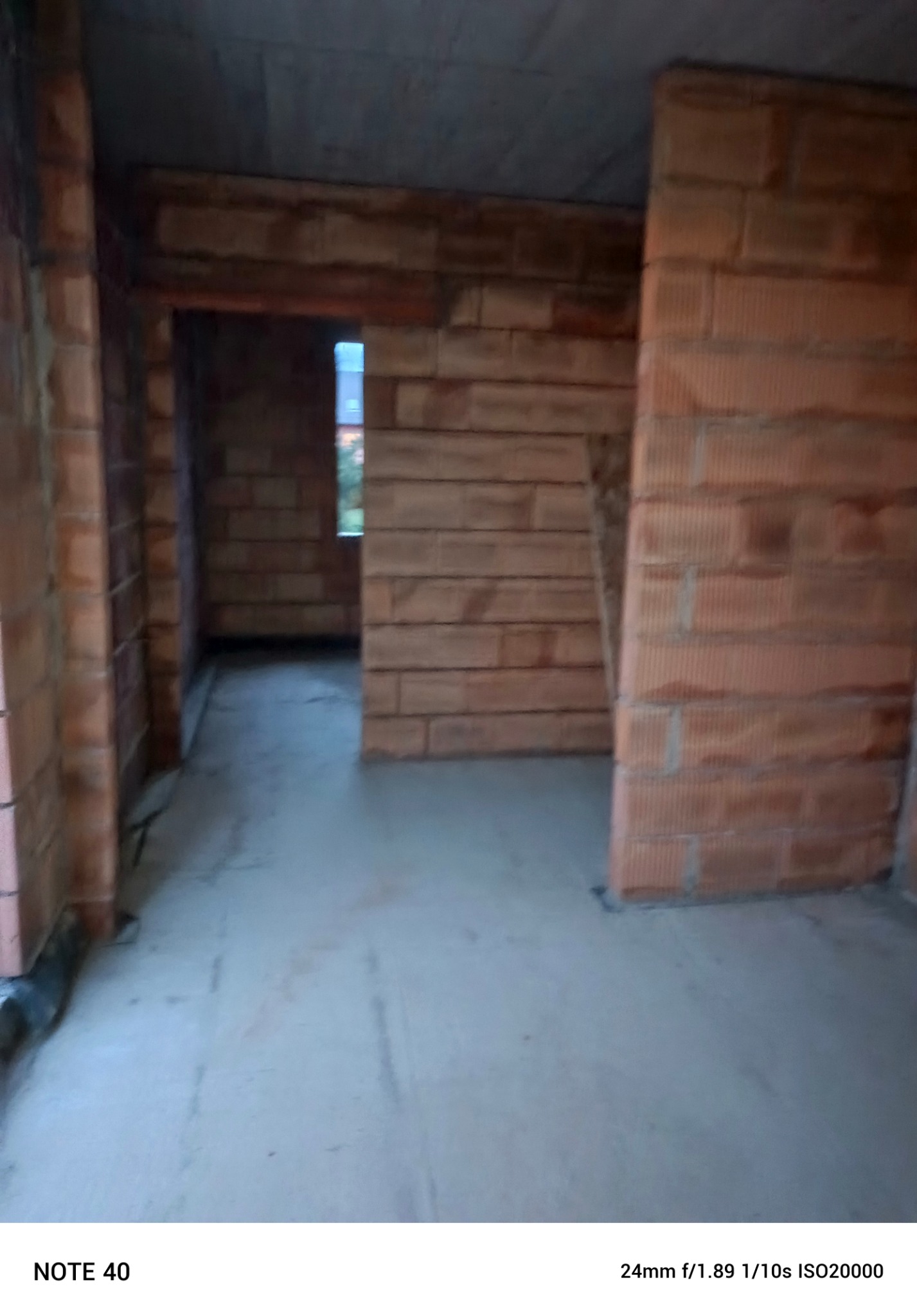 Surowe wnętrze w trakcie budowy z widocznymi ścianami z czerwonej cegły, niedokończonym betonowym sufitem i wylewką na podłodze, z oknem w tle.