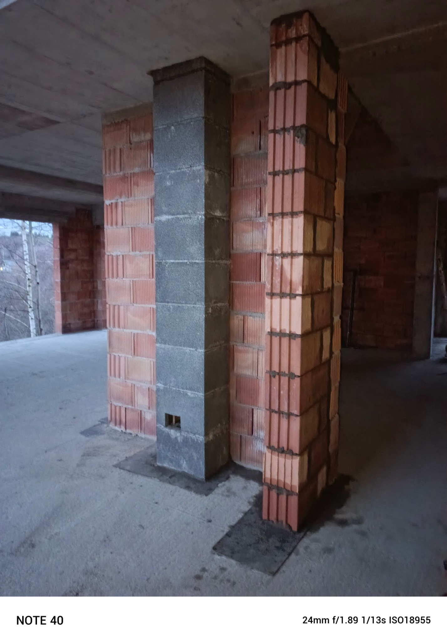 Wznoszenie pionowych słupów z czerwonej cegły dziurawki i szarego betonu komórkowego na surowym betonowym stropie.