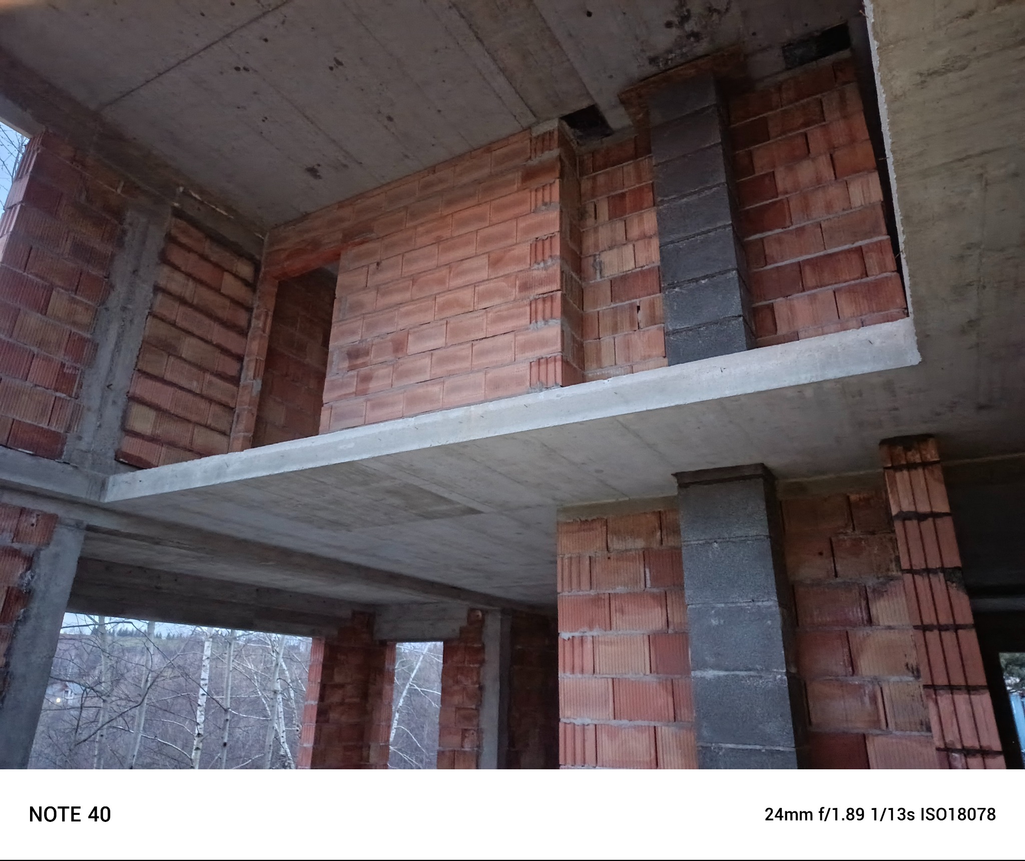 Surowy stan budynku w trakcie budowy: ściany z czerwonej cegły i szare betonowe filary, widoczne stropy, na zewnątrz drzewa bez liści w pochmurny dzień.