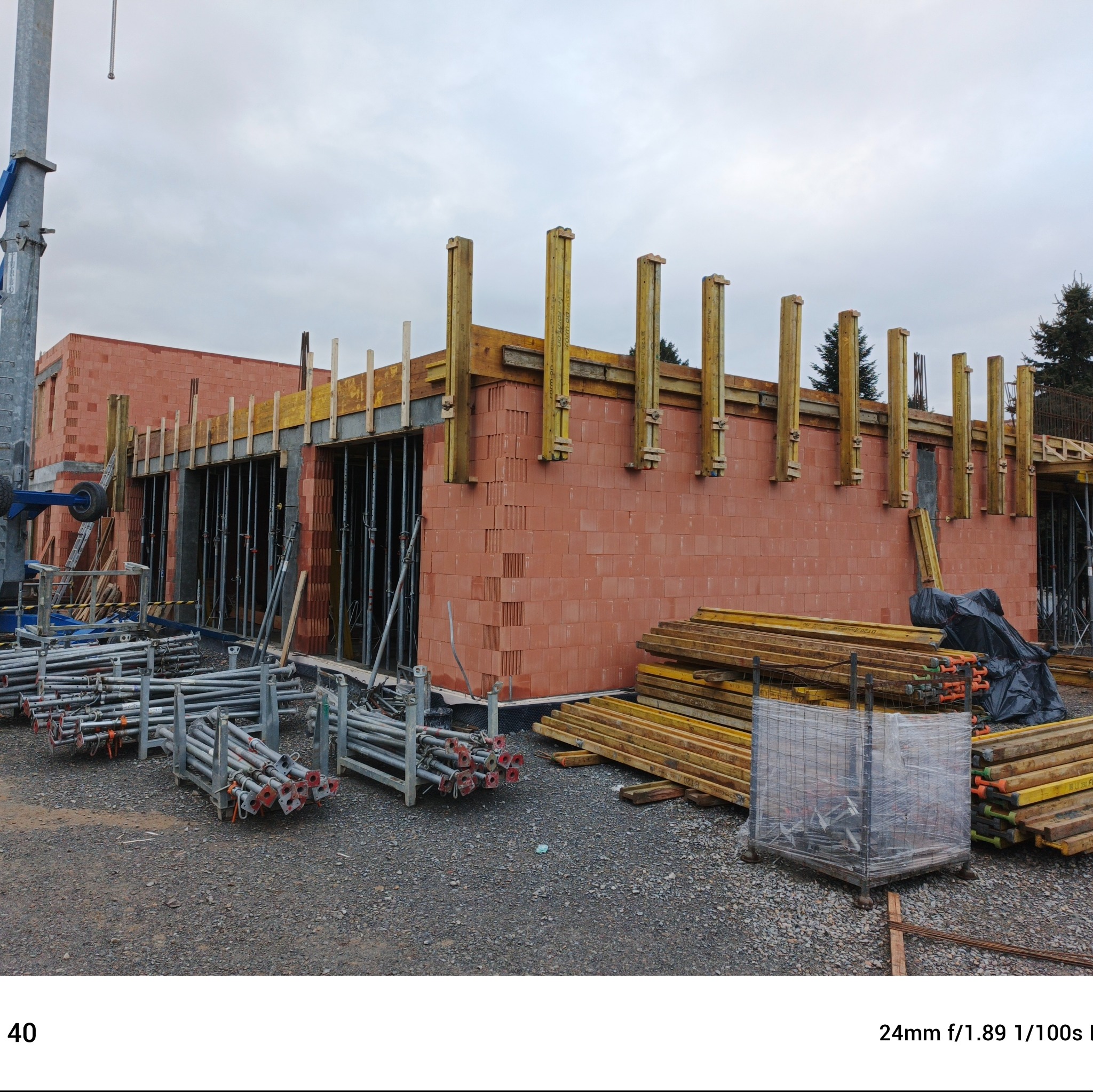 Murowana ściana budynku w trakcie budowy z widocznymi szalunkami i podporami stropowymi, na pierwszym planie rusztowania i materiały budowlane.