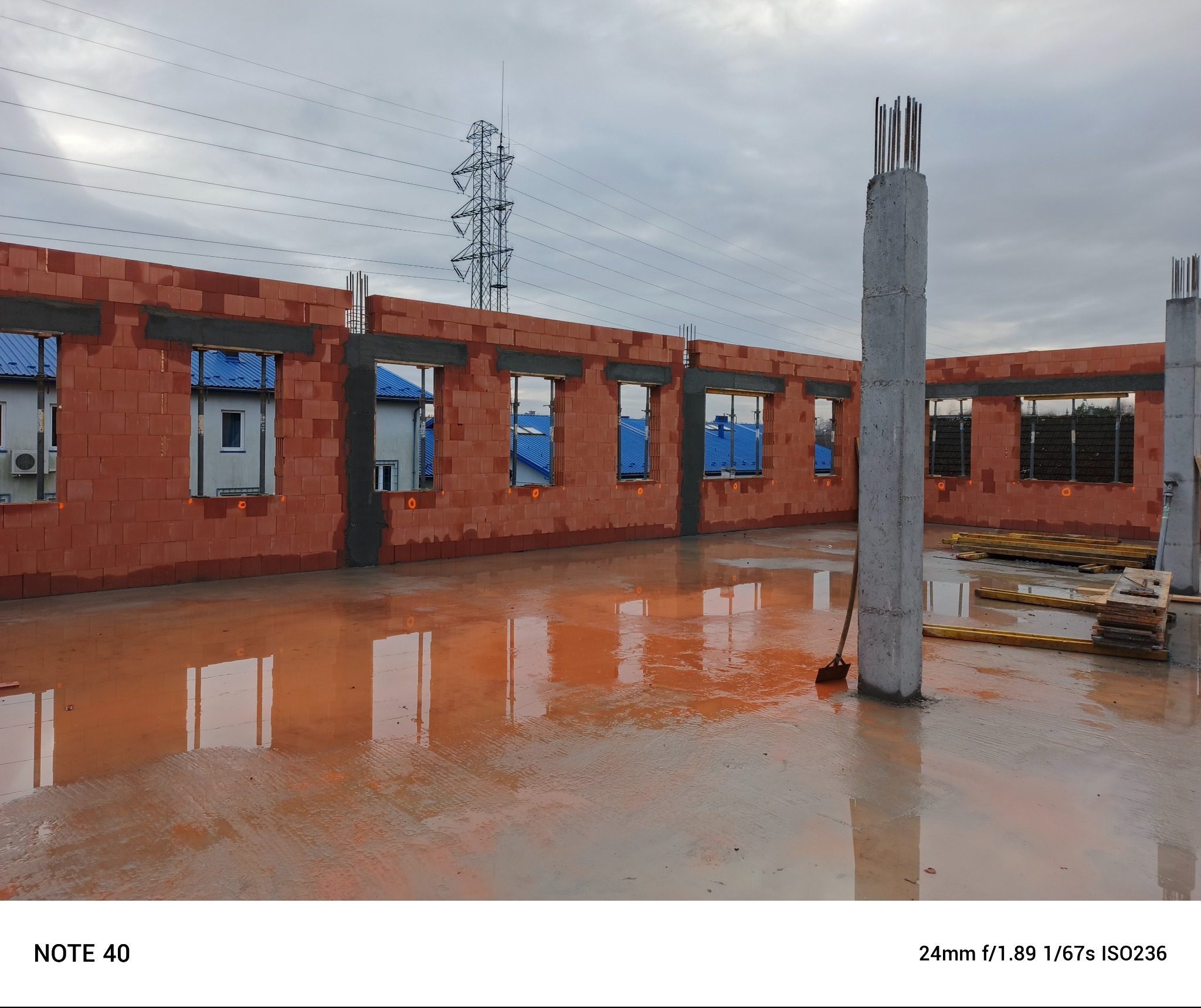 Wznoszone ściany z czerwonej cegły z otworami okiennymi, widoczne zbrojone słupy betonowe, mokra podłoga odbija elementy konstrukcji, w tle domy z niebieskimi dachami i słup wysokiego napięcia.