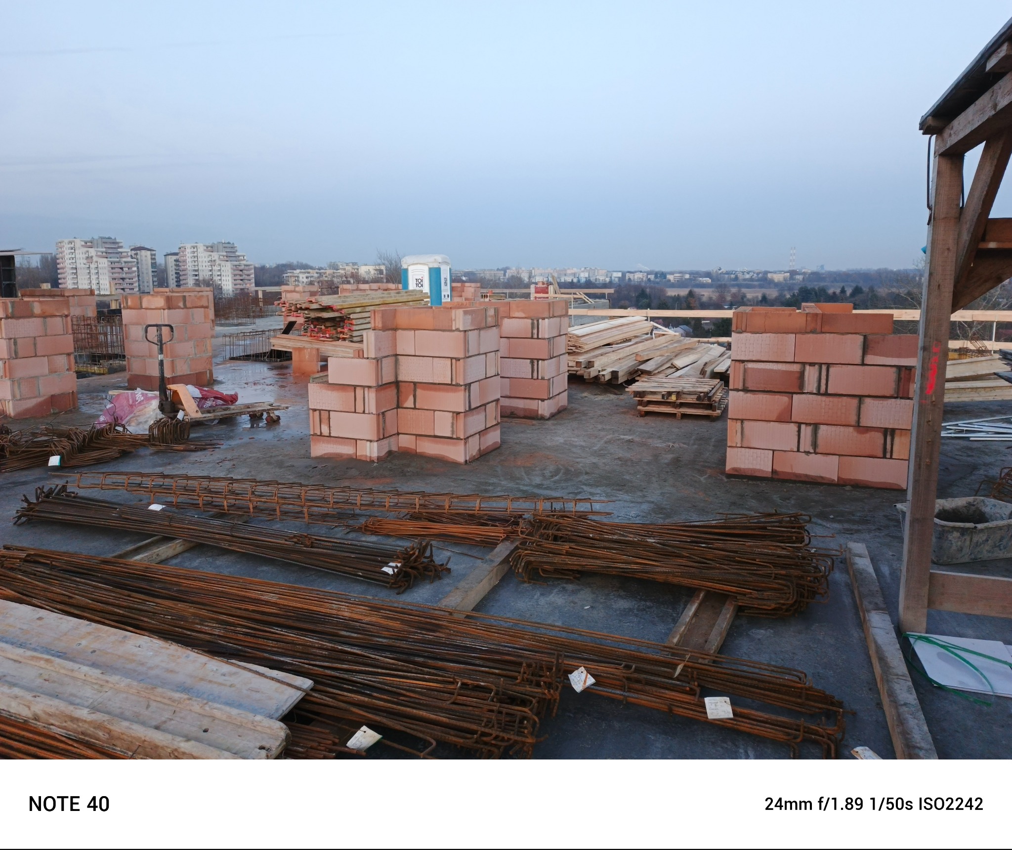 Wznoszone mury z czerwonej cegły na placu budowy, widoczne zbrojenia, drewniane elementy konstrukcyjne i panorama miasta w tle.