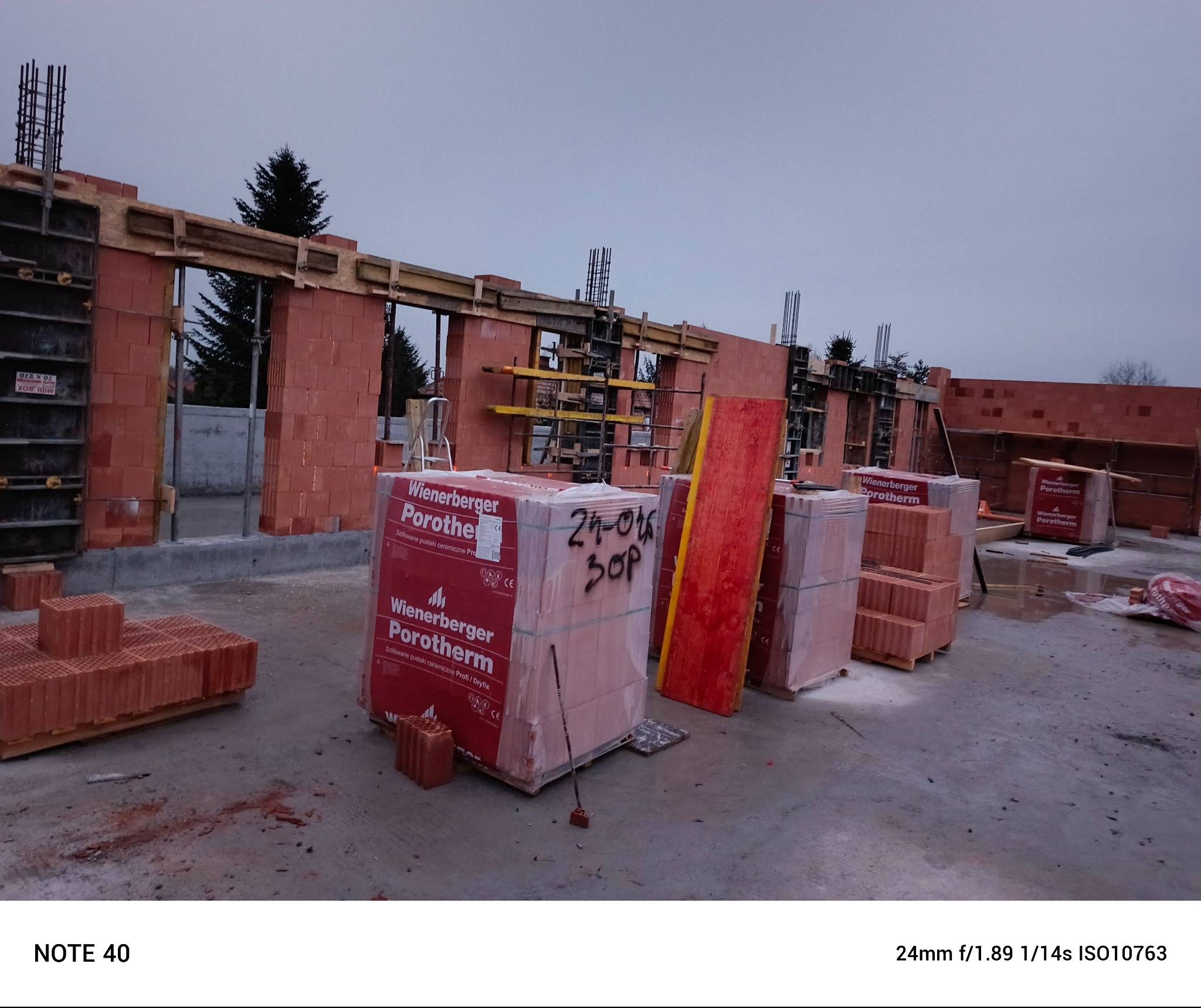 Budowa domu: wznoszenie ścian z czerwonej cegły Porotherm, widoczne szalunki i zbrojenia, na pierwszym planie opakowania cegieł z napisem 24-06 30P.