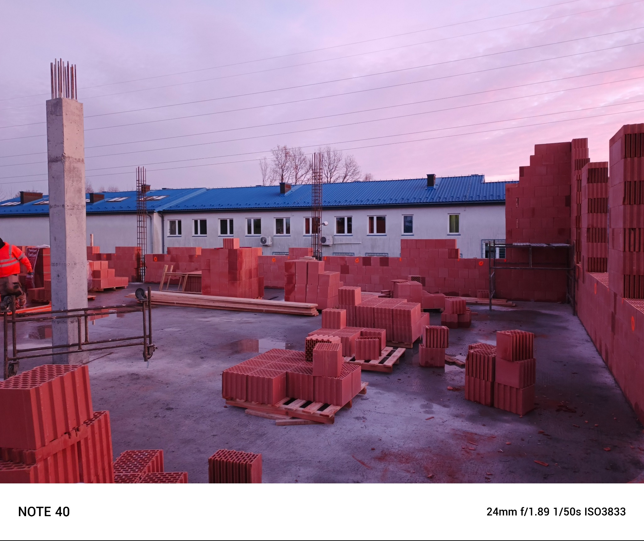 Wczesny etap budowy z czerwonej ceramiki poryzowanej: widoczne palety z bloczkami, fragmenty wzniesionych ścian oraz zbrojony słup na tle budynku z niebieskim dachem i różowego nieba.