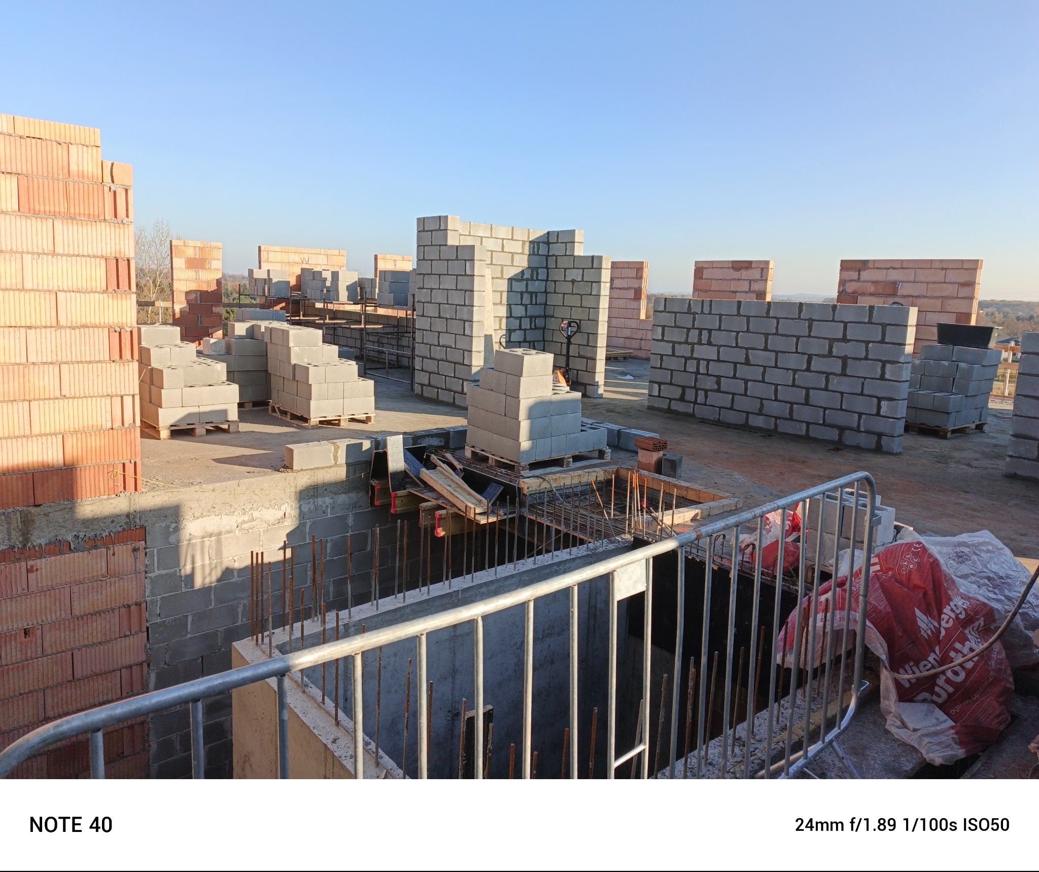 Wznoszone mury z bloczków i cegieł na budowie, widoczne zbrojenia i palety z materiałami budowlanymi w słoneczny dzień.