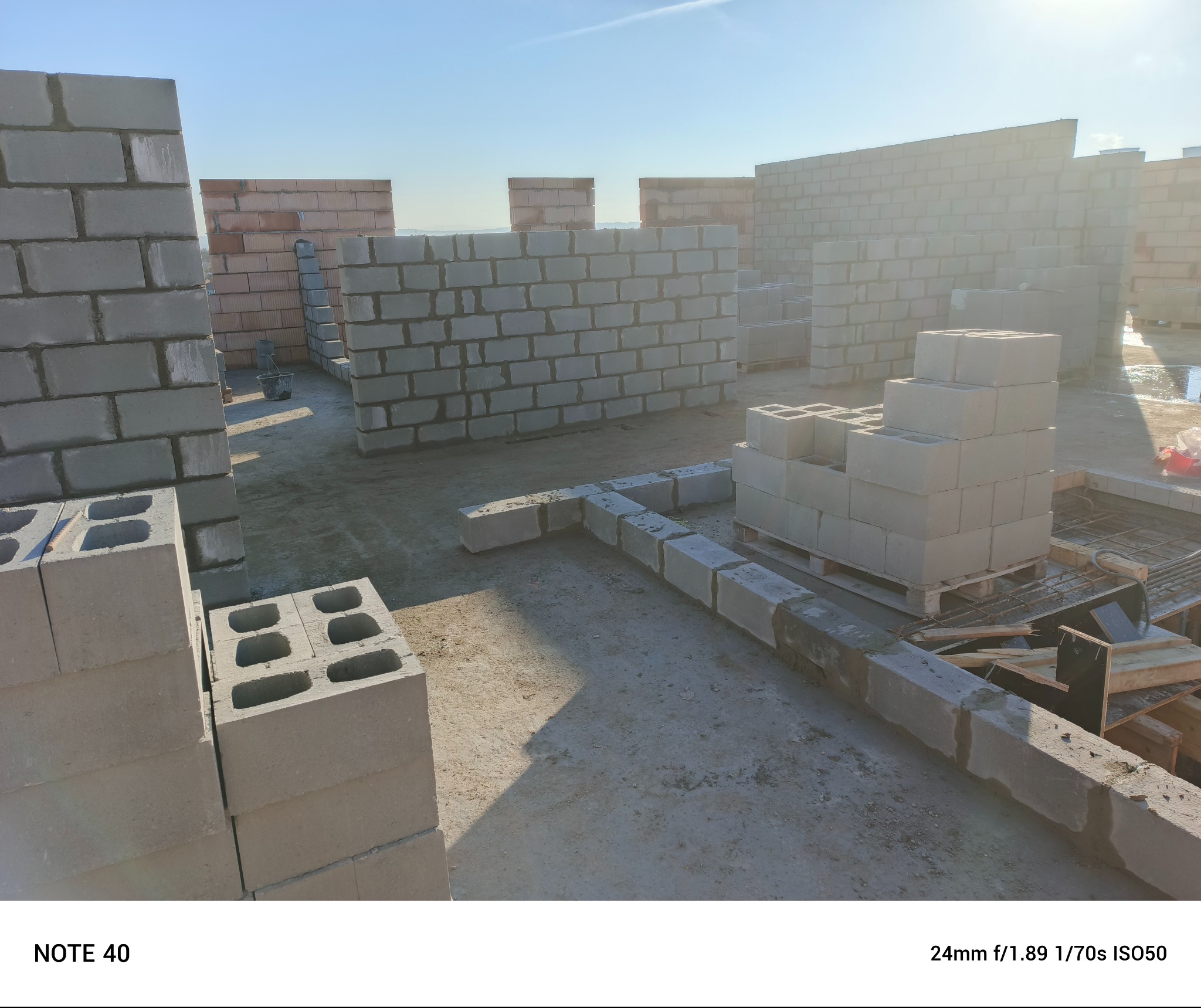 Wznoszone ściany z szarych bloczków betonowych na budowie, widoczne świeże spoiny i paleta z zapasem bloczków, jasne niebo w tle.