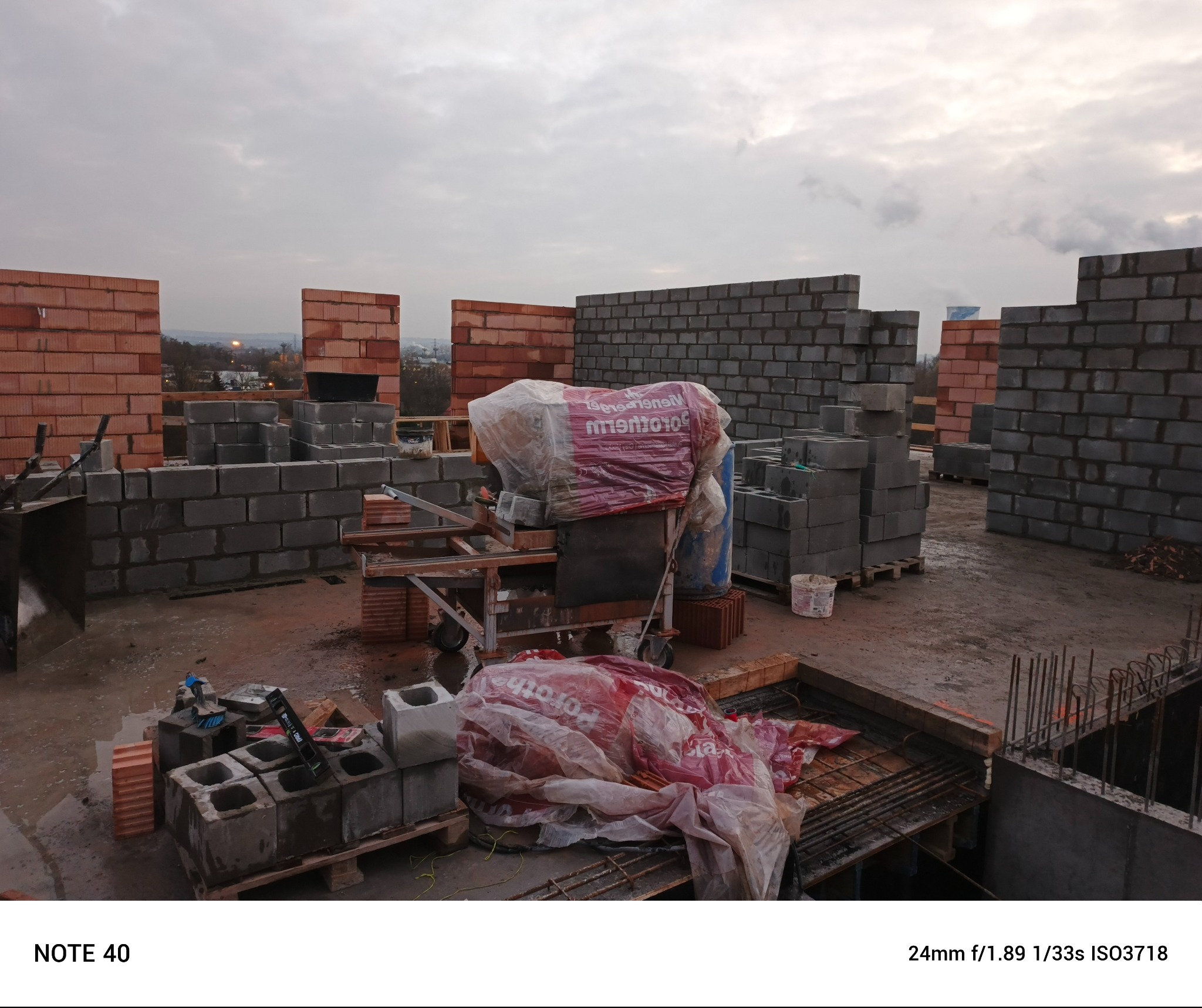 Wznoszenie murów z bloczków betonowych i cegieł Porotherm na budowie, widoczne narzędzia murarskie i materiały budowlane pod przykryciem folią, w tle zabudowa miejska pod pochmurnym niebem.