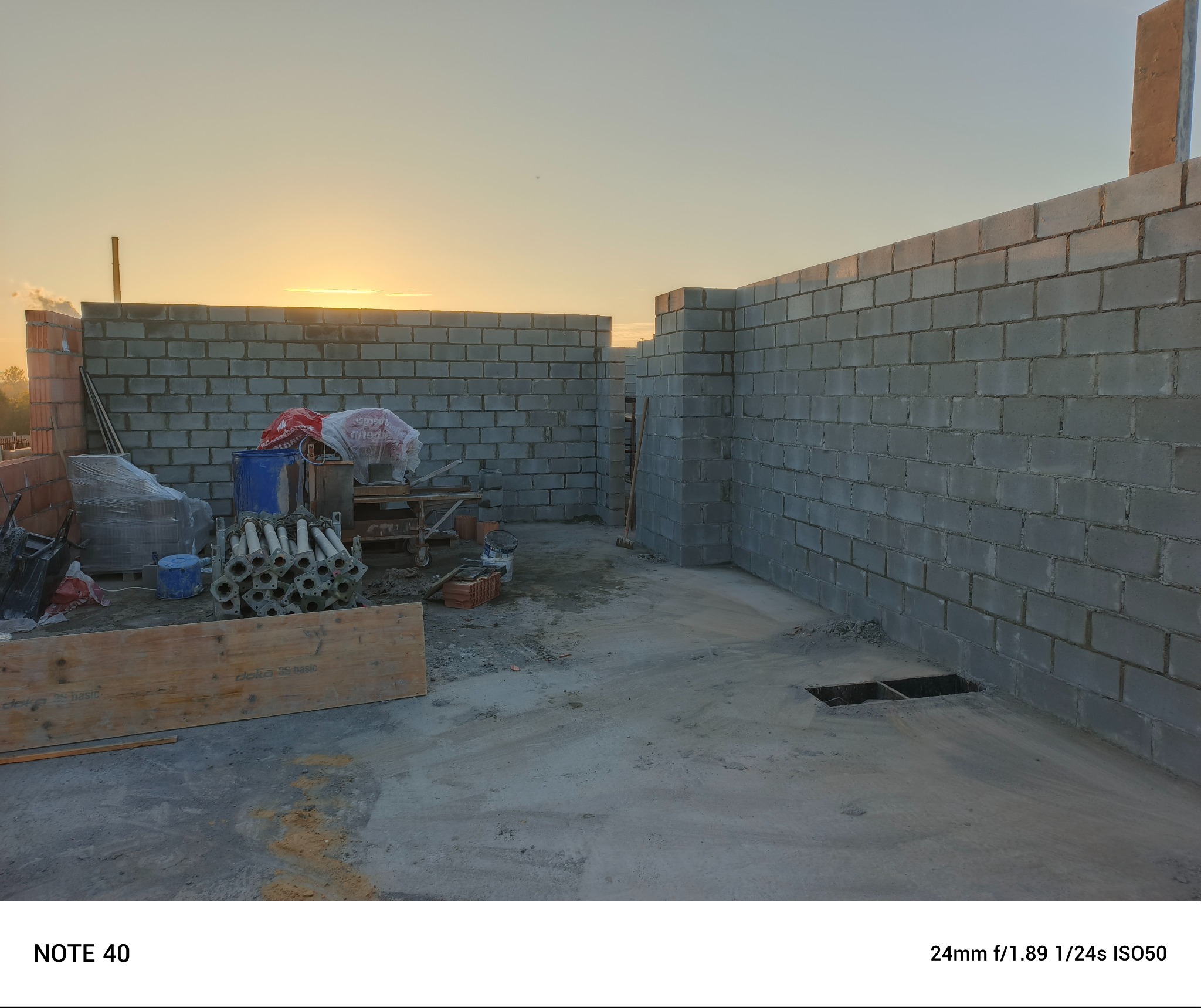 Wznoszone ściany z szarych bloczków betonowych na placu budowy, widoczne rusztowania, materiały budowlane i zachodzące słońce w tle.