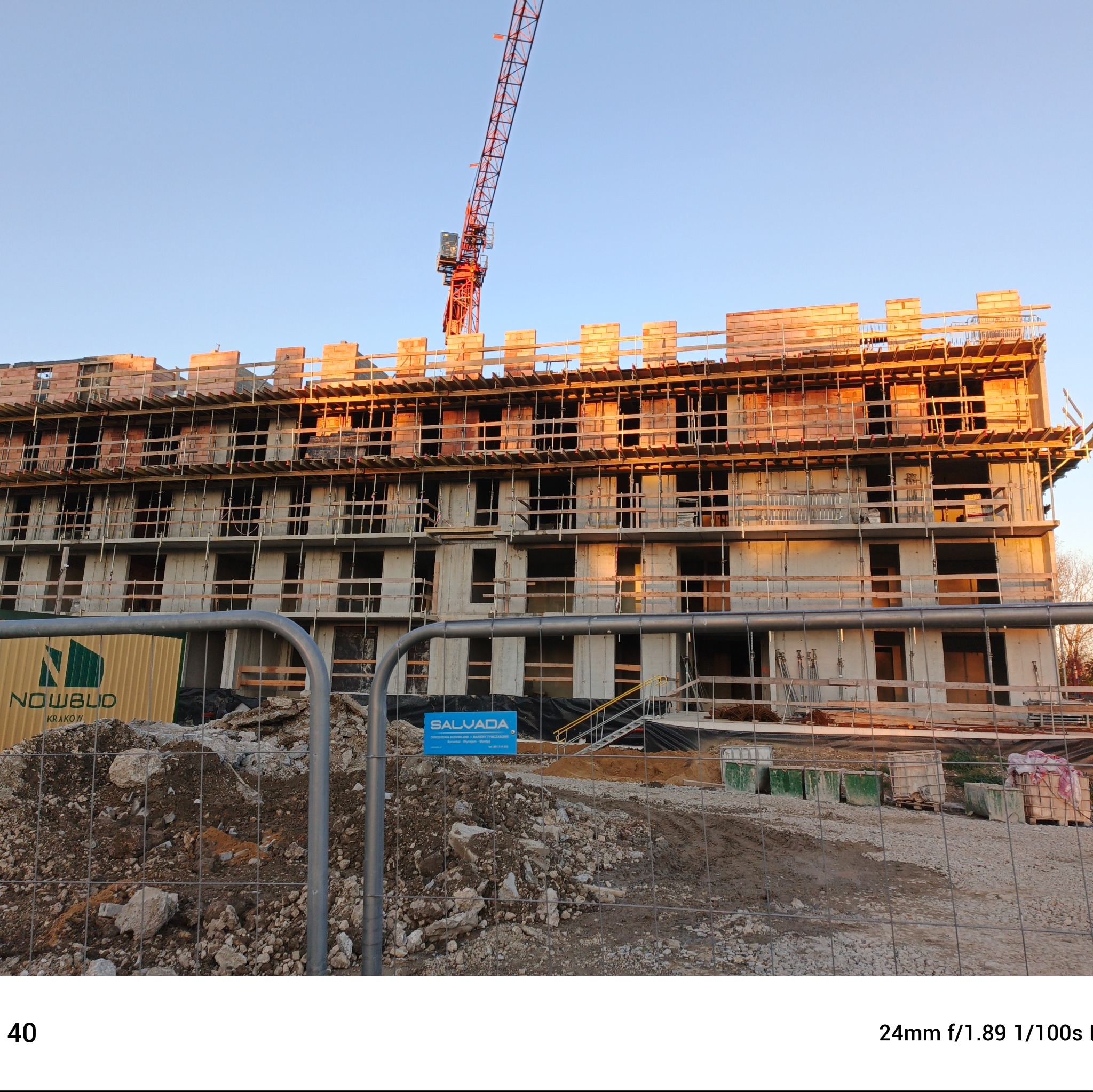 Wielopiętrowy budynek w trakcie wznoszenia, widoczne szare bloczki konstrukcyjne, drewniane szalunki i czerwony żuraw budowlany na tle błękitnego nieba, w dole ogrodzenie z siatki i gruz budowlany.