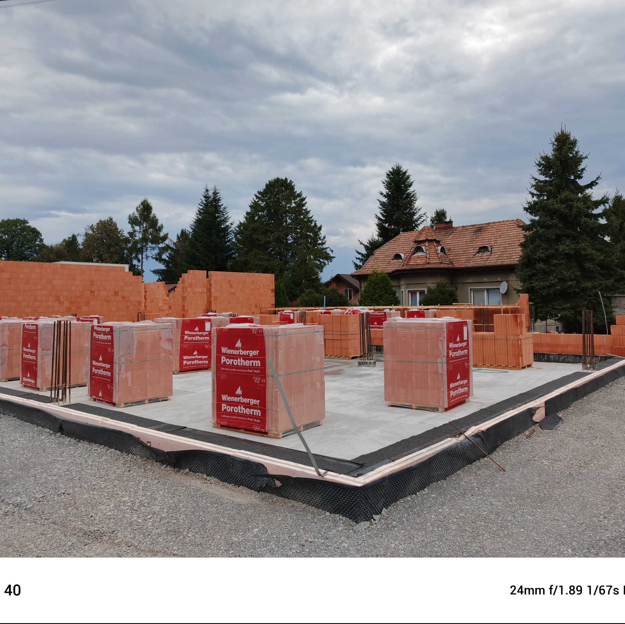 Fundamenty domu jednorodzinnego z widocznymi bloczkami Porotherm na paletach, przygotowane do wznoszenia ścian, w tle dom z czerwonym dachem i zielone drzewa.