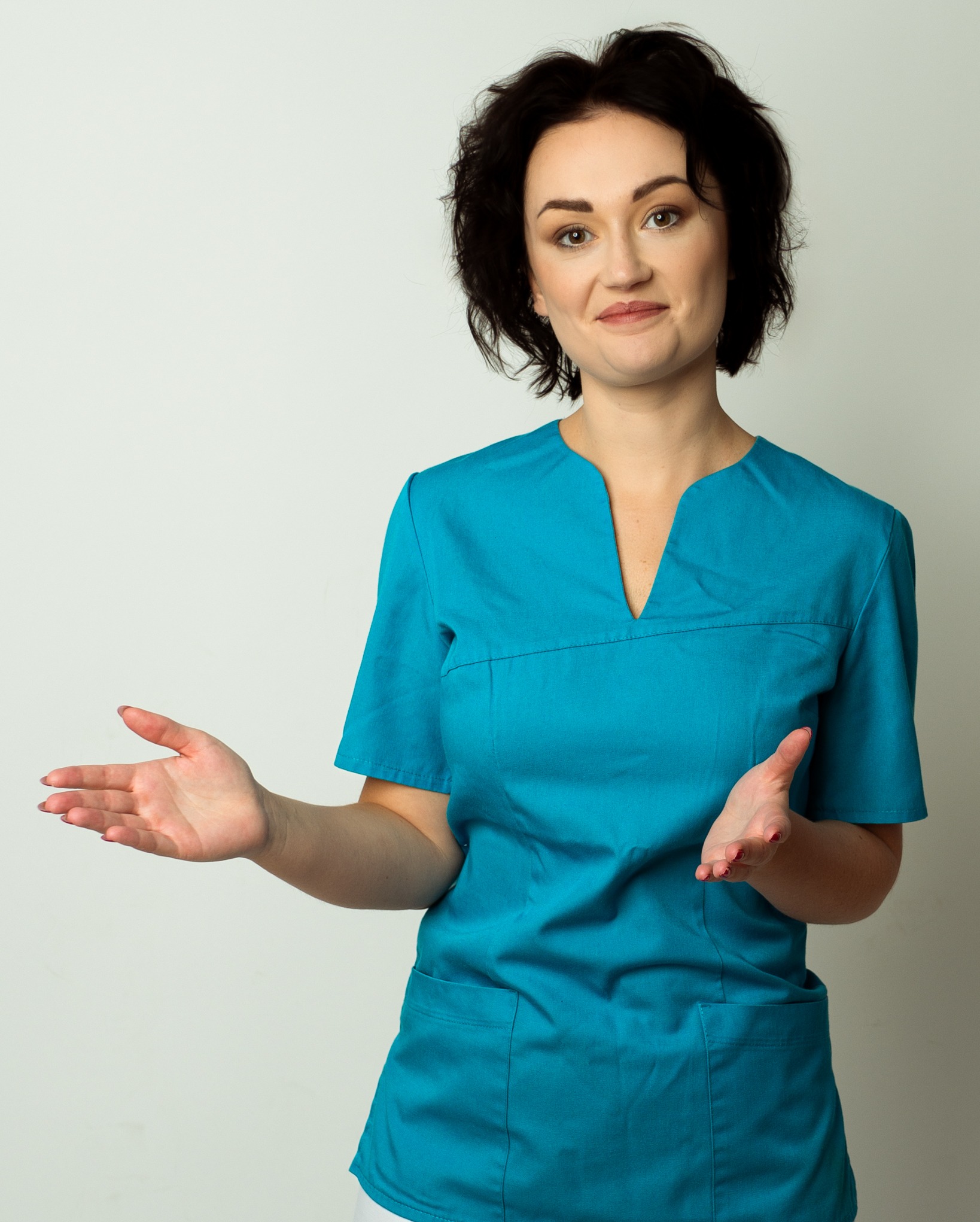 Uśmiechnięta kobieta w turkusowym uniformie prezentuje otwartymi dłońmi na jasnym tle. Krótkie, ciemne włosy, naturalny makijaż. Dwie kieszenie na uniformie.