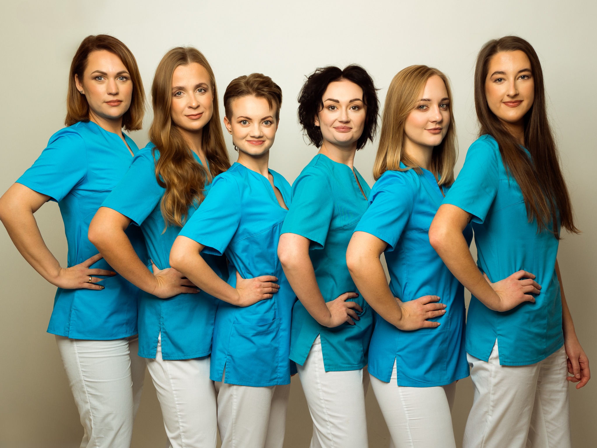 Zespół sześciu kobiet w turkusowych uniformach i białych spodniach, ustawionych w rzędzie, z rękami na biodrach, prezentuje się na jasnym tle.