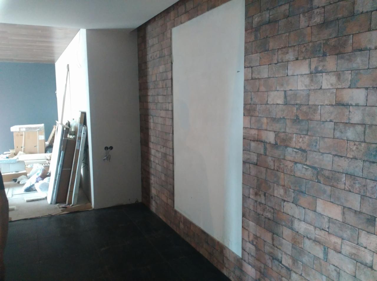 Wnętrze z fragmentem ściany wykończonej dekoracyjną cegłą, obok biała ściana i widoczne narzędzia oraz materiały budowlane.