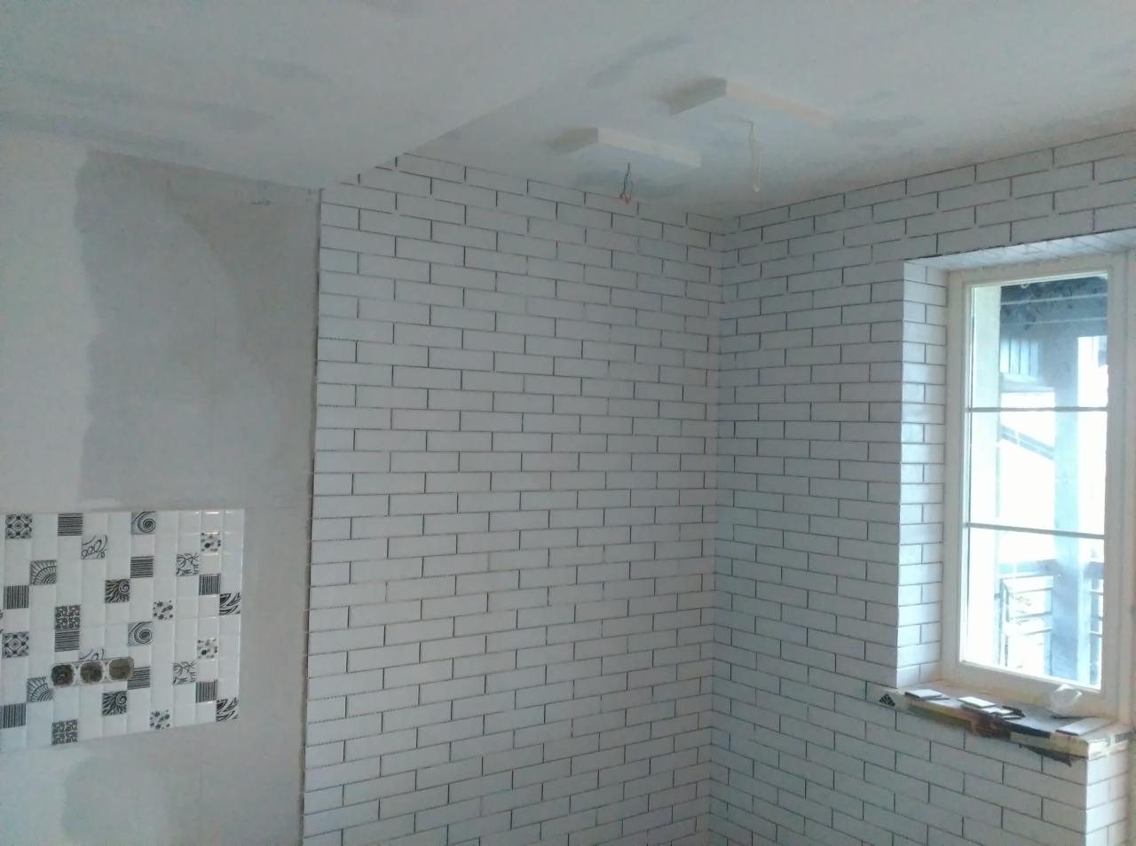 Ściana wyłożona białymi, podłużnymi płytkami ceramicznymi typu metro, z widocznym oknem i fragmentem ściany z dekoracyjnymi płytkami w czarno-białe wzory, sufit częściowo wykończony.