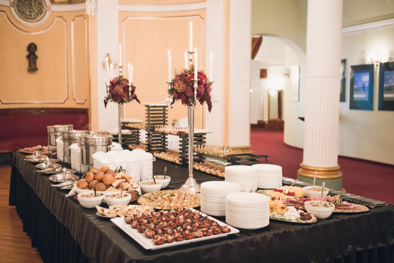 Elegancki stół cateringowy z różnorodnymi przekąskami, srebrnymi podgrzewaczami i świecznikami z kwiatami, ustawiony w klasycznym wnętrzu z kolumnami i czerwonym dywanem.