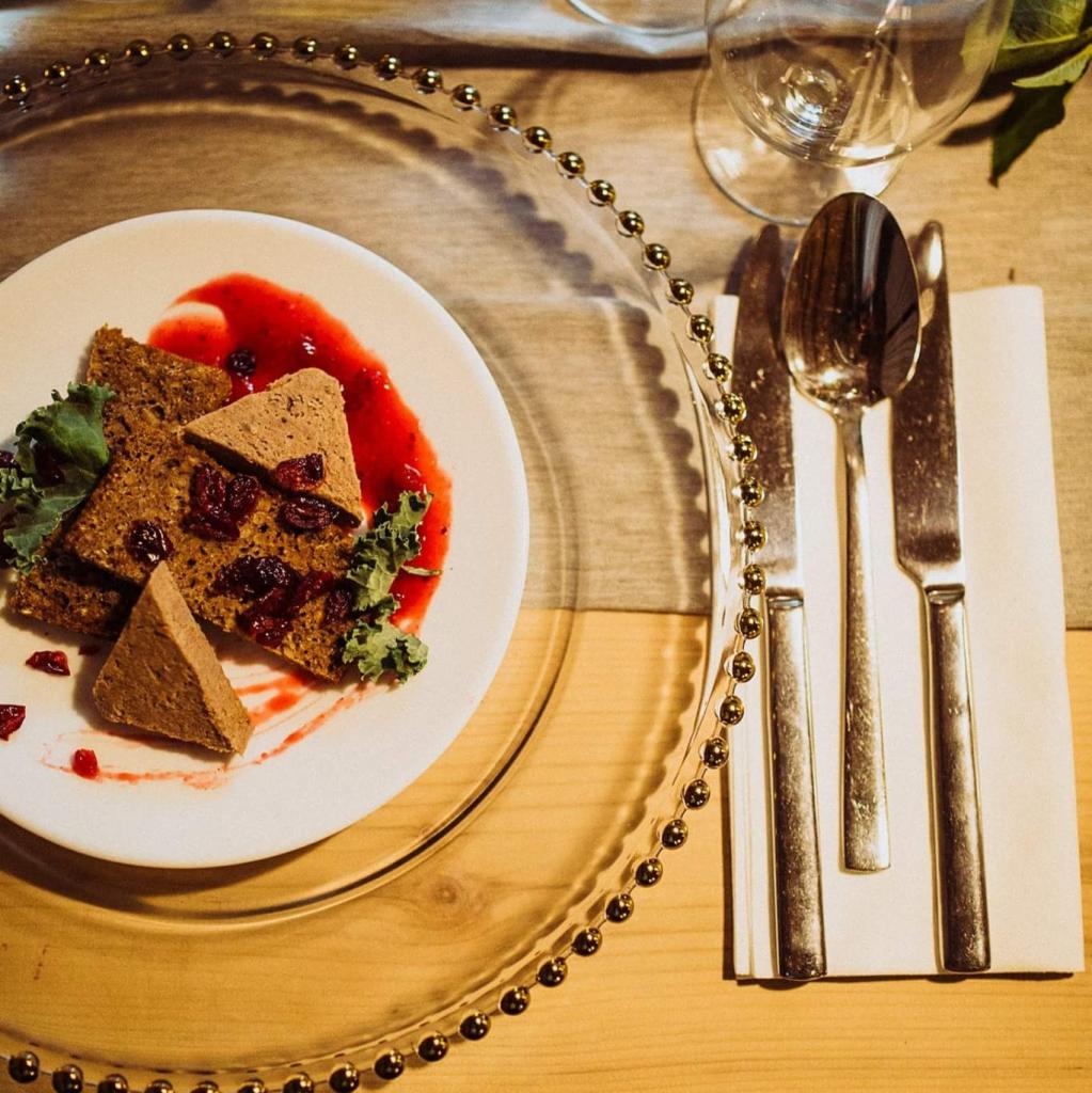 Elegancko podany pasztet z żurawiną na białym talerzu, udekorowany zielonymi listkami, polany czerwonym sosem. Talerz umieszczony na przezroczystej podkładce z dekoracją z koralików. Obok leżą...