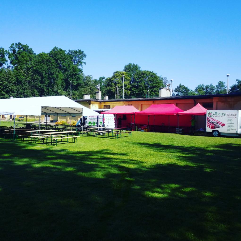 Rozstawione namioty cateringowe i stoły z ławkami na zielonej trawie, przygotowane do obsługi wydarzenia plenerowego w słoneczny dzień.