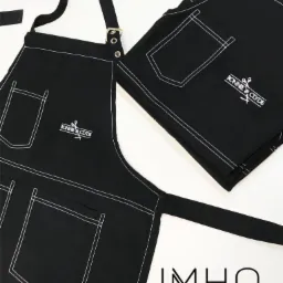 Dwa czarne fartuchy z białymi przeszyciami i logo pracowni krawieckiej, leżące na białym tle. Jeden fartuch rozłożony, drugi złożony w kostkę.