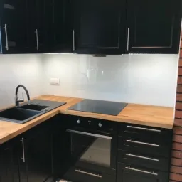 Nowoczesna kuchnia z czarnymi szafkami, drewnianym blatem i białym panelem szklanym nad blatem, oświetlonym listwą LED.