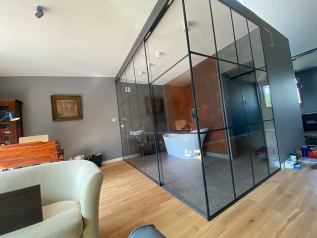 Szklana ścianka działowa z czarnymi profilami oddzielająca salon od łazienki z wolnostojącą wanną, widoczny fragment sofy i biurka w salonie.