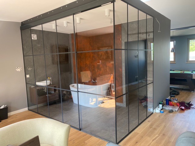 Szklana zabudowa w stylu industrialnym oddzielająca łazienkę z wolnostojącą wanną od reszty pomieszczenia, widoczny stół bilardowy w tle.