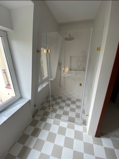 Szklana kabina prysznicowa z złotymi okuciami w łazience z oknem i biało-szarą podłogą w szachownicę.