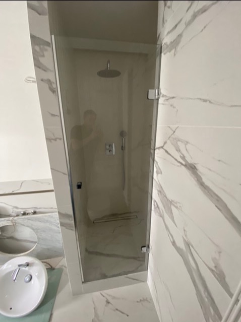 Kabina prysznicowa ze szklanymi drzwiami na zawiasach w łazience wykończonej białym marmurem, widoczna armatura i odpływ liniowy.