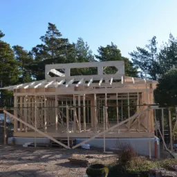 Surowy szkielet drewniany budynku w trakcie budowy, z widocznymi elementami konstrukcyjnymi i podporami, na tle lasu i błękitnego nieba.