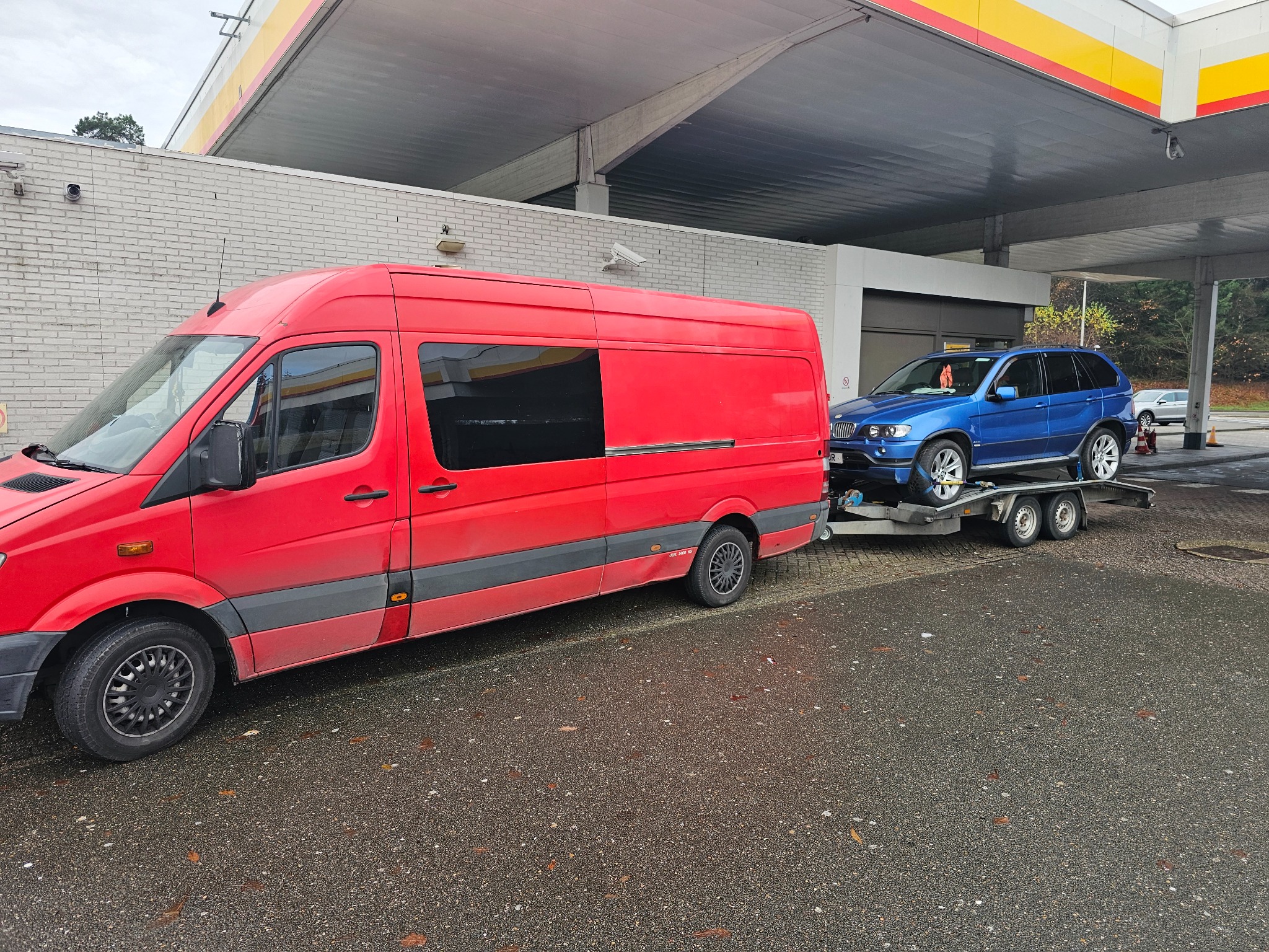 Czerwony bus transportowy z niebieskim samochodem osobowym na lawecie, zaparkowany na stacji benzynowej Shell.