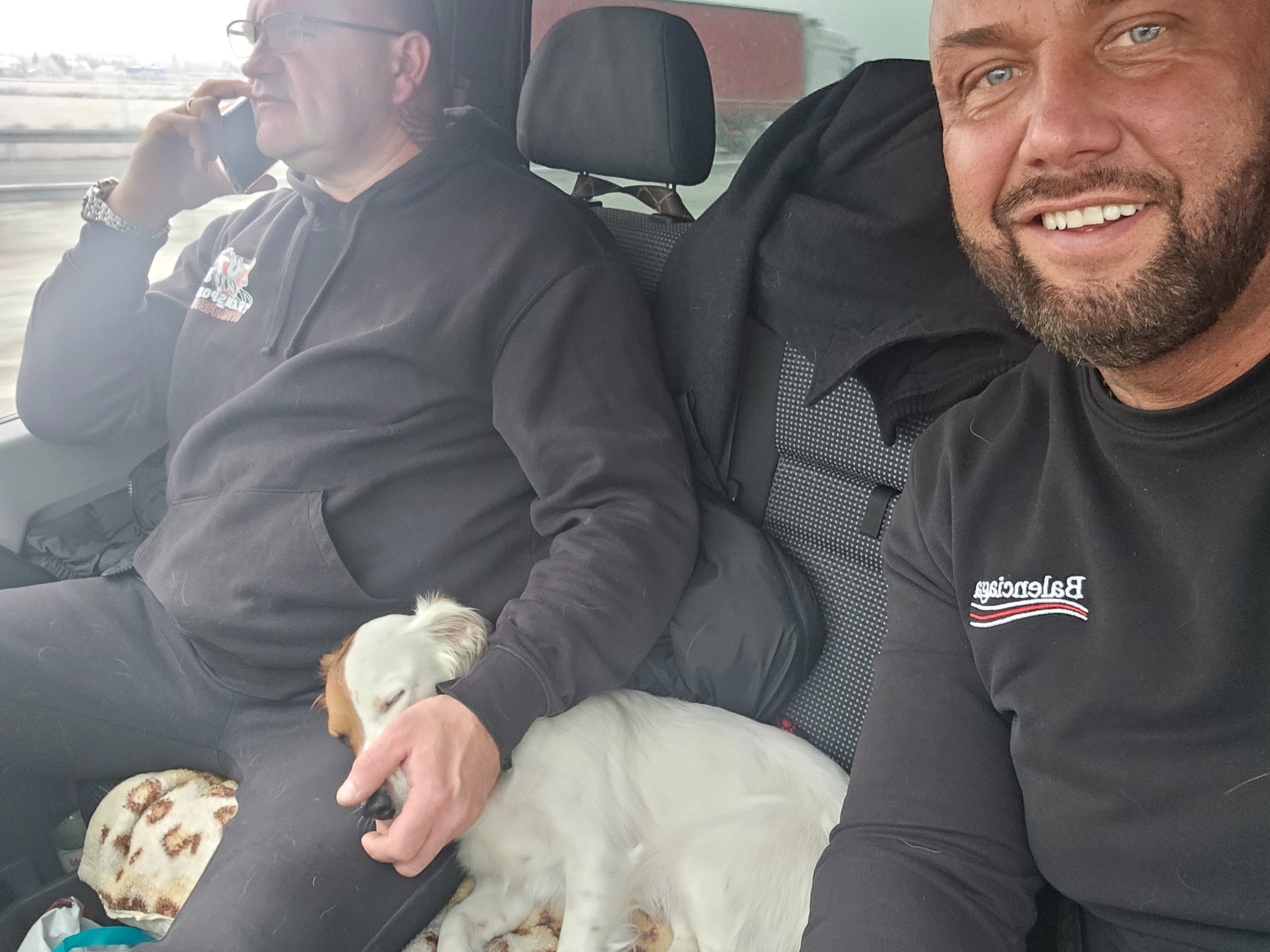 Dwóch mężczyzn podróżuje samochodem, jeden rozmawia przez telefon, drugi uśmiecha się do kamery, a na kolanach jednego z nich śpi pies.