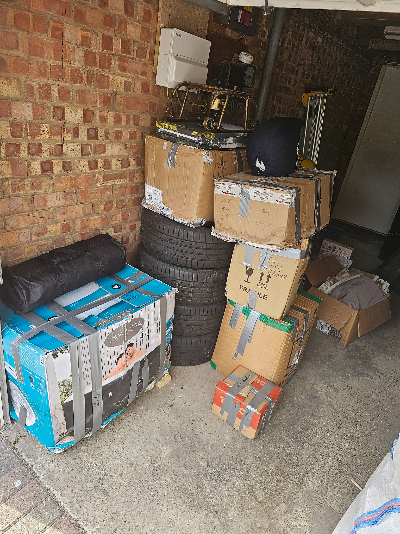 Stos kartonowych pudeł oklejonych taśmą, opon samochodowych i innych przedmiotów zgromadzonych w garażu, prawdopodobnie przygotowanych do transportu.