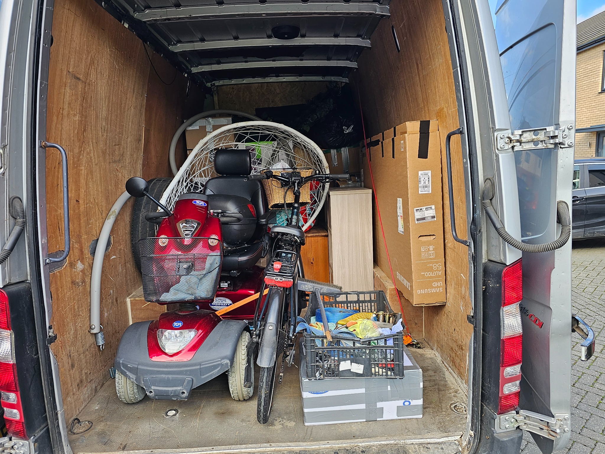 Wnętrze furgonetki załadowane różnymi przedmiotami: skuter inwalidzki, rower z wiklinowym koszykiem, krzesło wiszące, kartony i skrzynka z narzędziami, przygotowane do transportu.