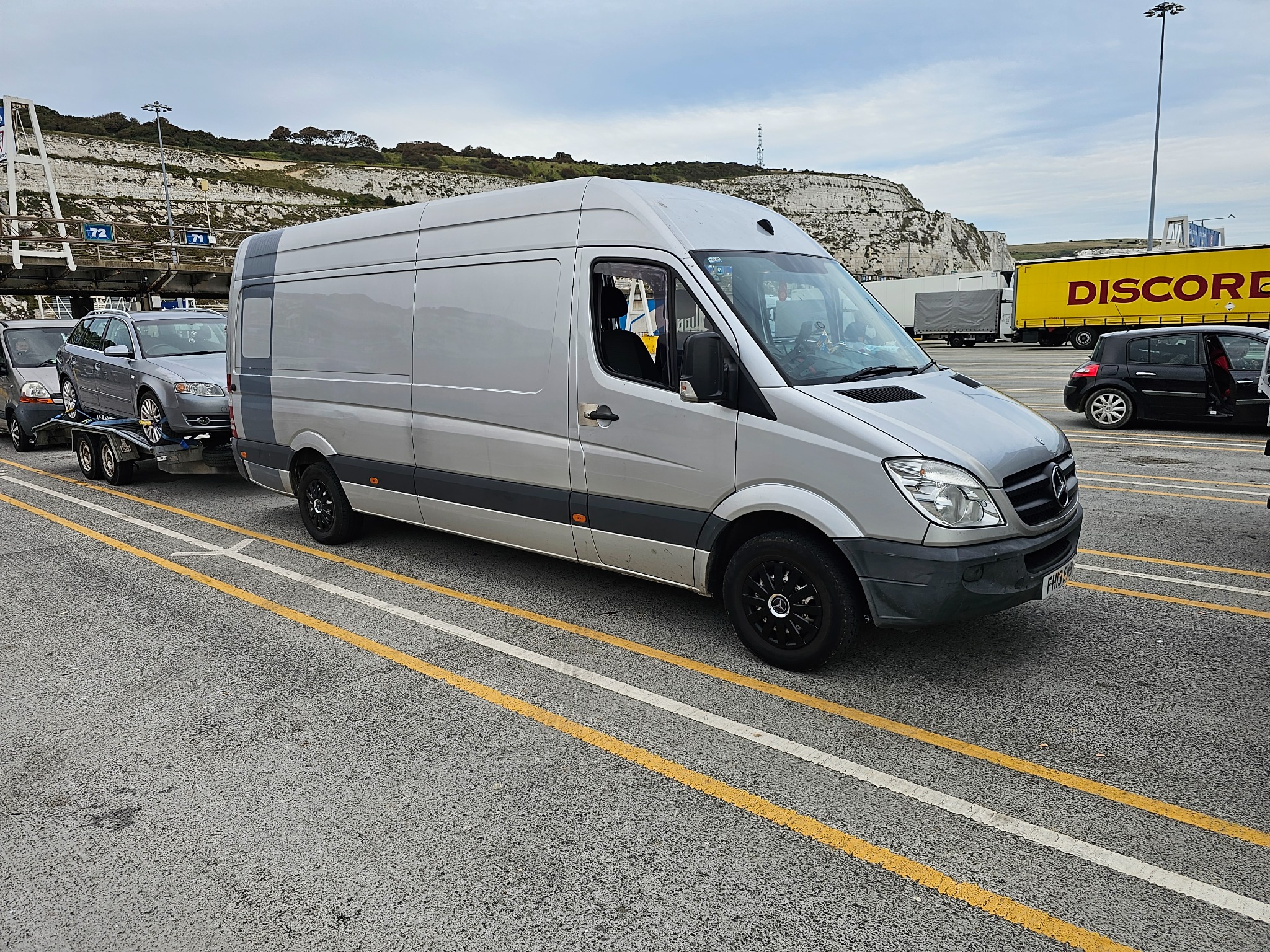Srebrny van z przyczepą transportującą srebrny samochód osobowy, stoi na pasie w oczekiwaniu na prom, w tle białe klify Dover.