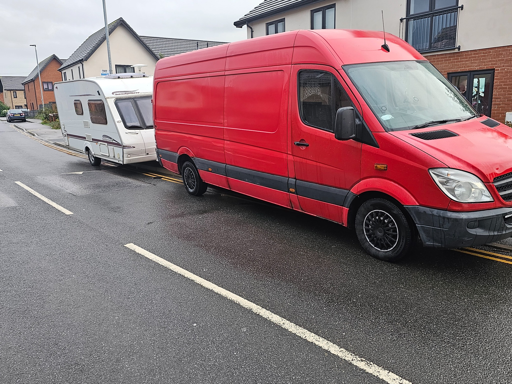 Czerwony van holowniczy z białą przyczepą kempingową na mokrej ulicy w dzielnicy mieszkalnej.