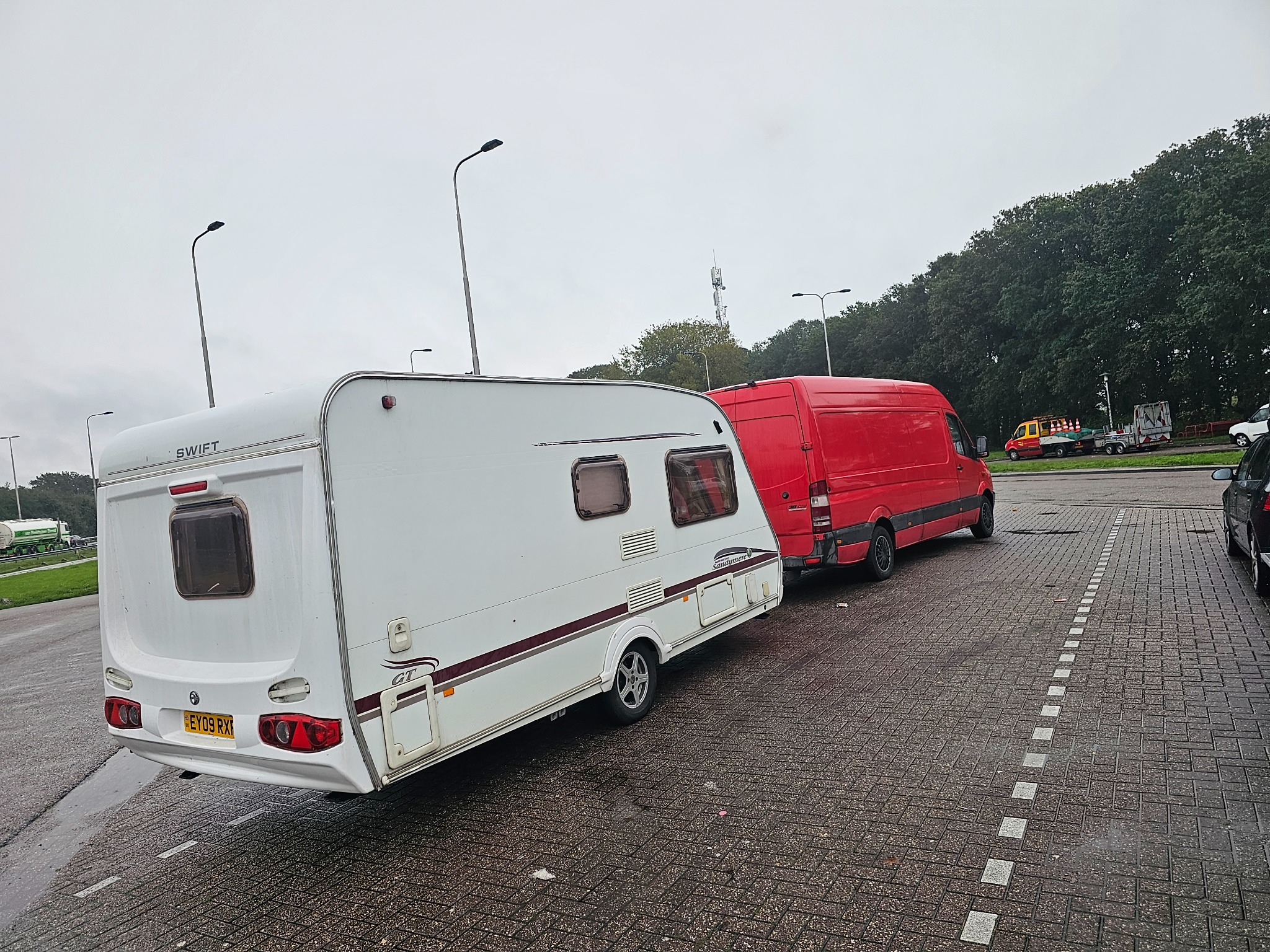 Biała przyczepa kempingowa Swift GT holowana przez czerwony samochód dostawczy na parkingu z kostki brukowej w pochmurny dzień.