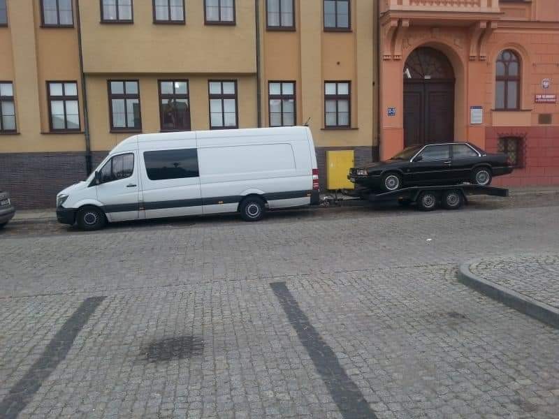 Biały bus z przyczepą transportującą czarny samochód osobowy na tle budynku w mieście.
