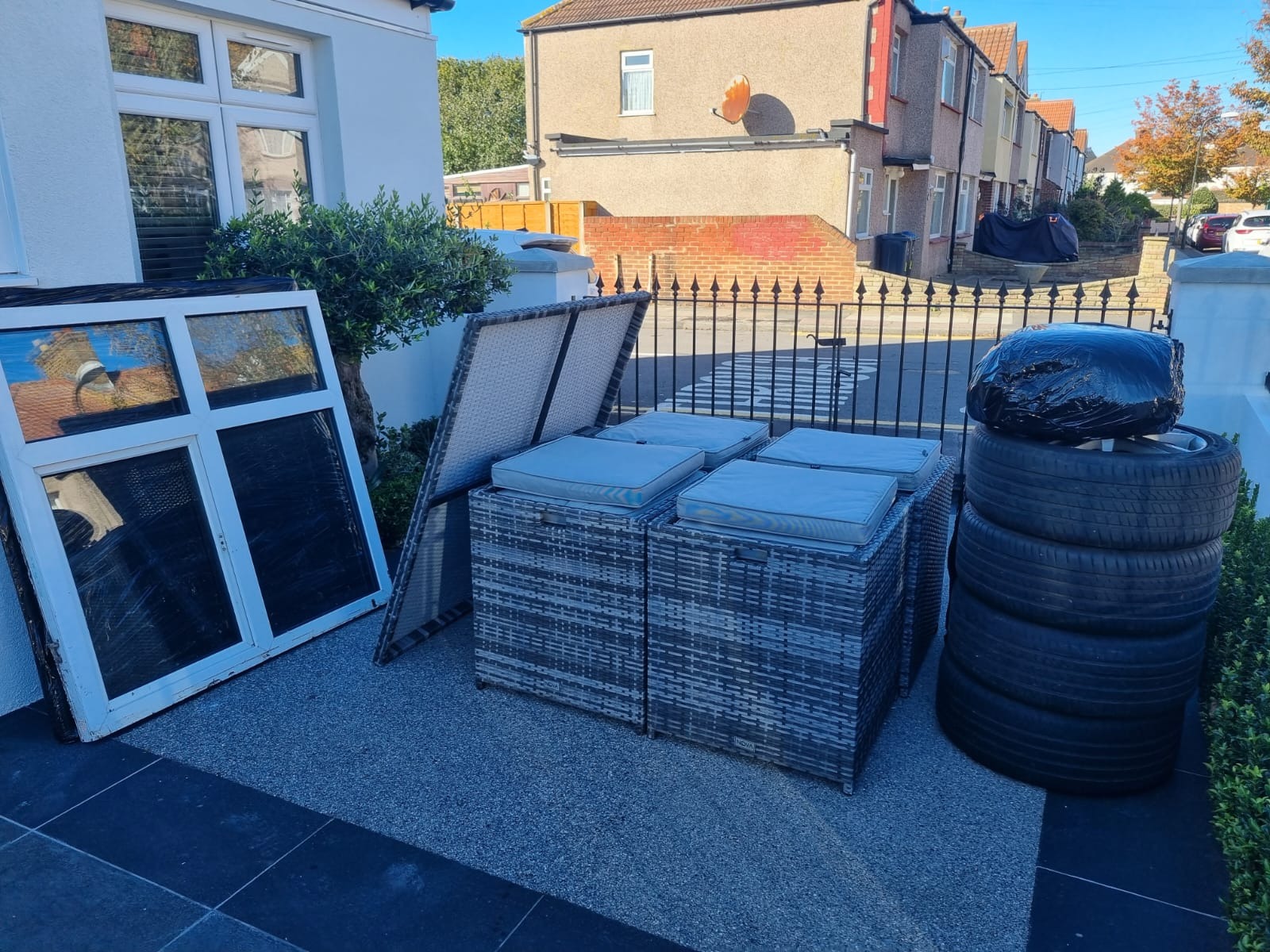 Meble ogrodowe z poduszkami, okno w ramie i opony w czarnym worku przygotowane do transportu przed domem.