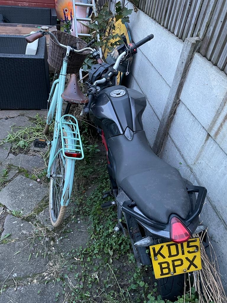 Niebieski rower z wiklinowym koszykiem i szary motocykl zaparkowane obok betonowego ogrodzenia, widoczne trawa i chwasty.