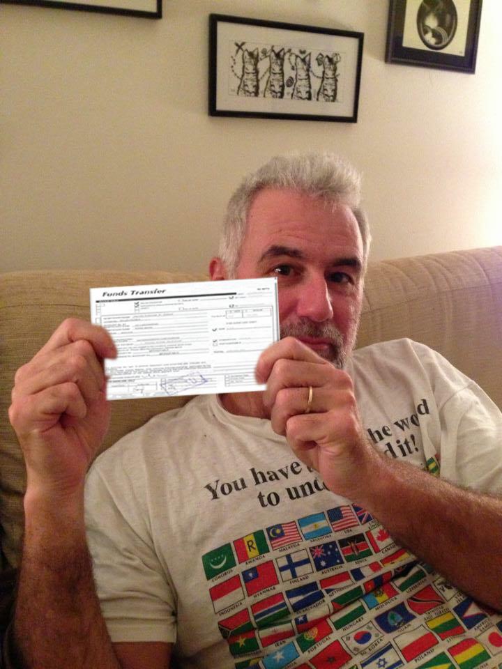 Mężczyzna z siwymi włosami i brodą siedzący na kanapie, trzymający w dłoniach formularz przelewu środków; w tle ściana z obrazkami.