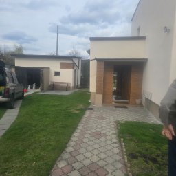 MALBUD - Nowoczesny dom jednorodzinny z garażem, jasna elewacja, drewniane akcenty przy wejściu, kostka brukowa na podjeździe, trawnik. Widok z perspektywy ogrodu.