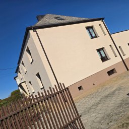 MALBUD - Jasna elewacja domu z brązowymi akcentami, widoczna zza brązowego ogrodzenia. Błękitne niebo w tle. Ukośne ujęcie podkreśla geometrię budynku.