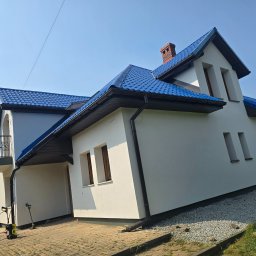 MALBUD - Biały dom z niebieskim dachem po remoncie elewacji, widok z dołu. Pracownik z myjką ciśnieniową w tle. Uwagę przyciąga intensywny kolor dachu.
