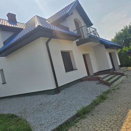 MALBUD - Biały dom z niebieskim dachem i balkonem z kutą balustradą, widok z dołu, kostka brukowa na podjeździe, drzewa w tle, słoneczny dzień.